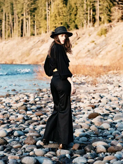 A woman in flowing black attire and wide-brimmed hat stands contemplatively among smooth river stones along the Floodplain Trail in Multnomah County, Oregon. Golden hour light bathes the scene, casting warm tones across the pebbled riverbed while dense forest rises in the soft-focused background. Her pose suggests quiet introspection as she gazes across the water, creating an atmospheric portrait of solitude in nature.