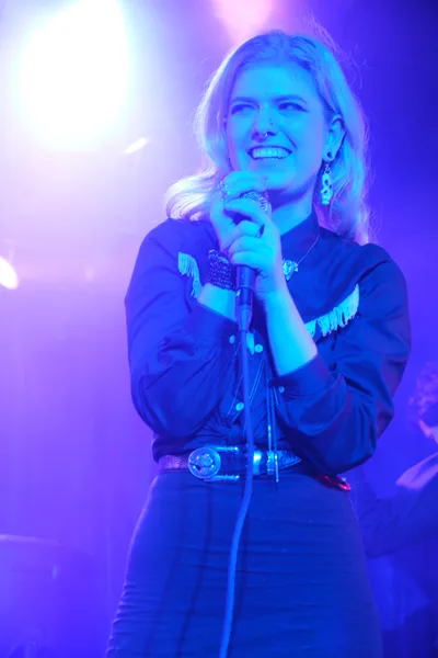 Faith Grossnicklaus of Roselit Bone grips her microphone with both hands, her blonde hair catching the electric blue and purple stage lighting at Dante's club in Portland. The intimate venue's atmospheric lighting transforms the performance space into a dreamy wash of cyan and violet hues, creating an otherworldly backdrop for the violinist's captivating stage presence. Her western-inspired shirt with fringe details and ornate belt buckle blend country aesthetics with the club's moody ambiance.