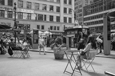 In the bustling heart of Herald Square, Manhattan, pedestrians claim moments of urban tranquility at scattered café tables amid the commercial chaos. The monochromatic composition captures the rhythmic dance of city life, where solitary figures pause with coffee and conversation against a backdrop of towering brick facades and Korean signage. Geometric folding chairs and planters create intimate pockets of respite, while streams of shoppers navigate between the outdoor seating and storefront displays.