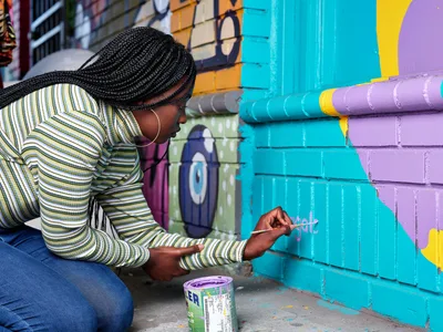 Naomi Likayi, wearing a striped long-sleeve shirt and gold hoop earrings, carefully signs her completed mural on Northeast 15th Avenue in Portland's Alberta Arts District. The artist kneels on the concrete sidewalk, paintbrush in hand, adding her signature to vibrant turquoise and purple geometric shapes that transform the brick wall. Behind her, the colorful street art creates a dynamic backdrop of urban creativity in this historically significant neighborhood.