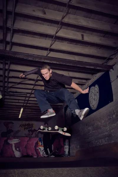 A young skater crouches in focused concentration within the gritty basement confines of Commonwealth Skateboarding in Portland's Buckman neighborhood. The weathered concrete walls and exposed ceiling beams create a moody industrial backdrop, while graffiti and skateboard graphics add splashes of color to the underground space. The atmospheric lighting captures the raw authenticity of Portland's skate culture, with the subject positioned dynamically against the textured urban environment.