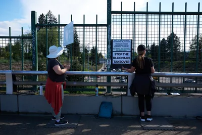 Two women stand at a concrete barrier along North Skidmore Street in Portland's Humboldt neighborhood, positioned above Interstate 5 for a May Day banner drop. One activist wears a white sun hat and red skirt, while her companion in dark clothing leans against the railing. Behind them, protest signs reading "STOP THE BILLIONAIRE TAKEOVER" are mounted on green security fencing, creating a scene of grassroots resistance under the Pacific Northwest's characteristic overcast sky.