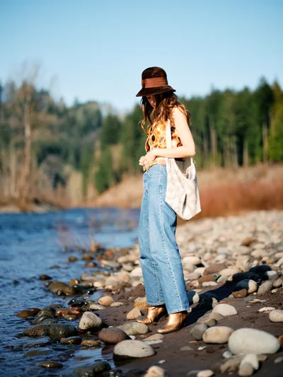 A young woman in vintage-inspired attire stands contemplatively on the rocky shoreline of the Columbia River along Multnomah County's Floodplain Trail. Wearing a distinctive brown felt hat, sunflower-print top, and high-waisted denim, she gazes downward while holding a canvas tote bag. The golden hour light illuminates her figure against the serene water and autumn-touched forest backdrop, creating an intimate moment of reflection in Oregon's natural landscape.