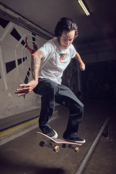 A tattooed skateboarder navigates through the industrial passage of Commonwealth Skateboarding in Portland's Buckman neighborhood. Fluorescent lighting casts stark shadows across concrete walls as the rider demonstrates fluid motion on weathered pavement. The subterranean tunnel creates an intimate urban skating environment, with white structural elements and yellow safety markings defining the confined space.