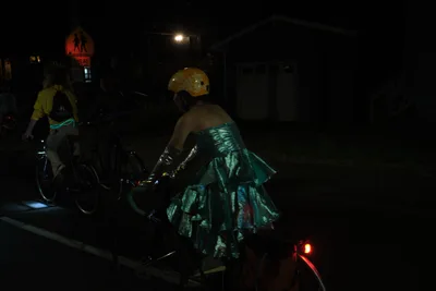 Under the cover of Portland's Richmond neighborhood darkness, two cyclists navigate the streets in an enchanting juxtaposition of formal elegance and urban mobility. The foreground rider wears a lustrous teal gown that catches the sparse streetlight, creating ethereal ripples of fabric against the night. A golden helmet provides a striking contrast to the flowing dress, while warning signs and distant street lamps punctuate the mysterious nocturnal scene.
