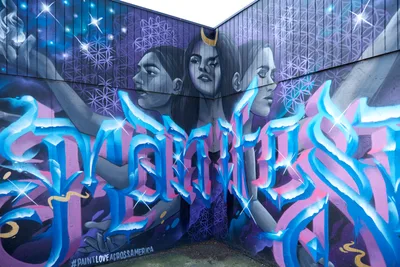 Three ethereal faces crowned with golden crescents gaze from a corner mural in Portland's Central Eastside, their monochromatic features floating above an explosive wildstyle composition in electric blues and magentas. Sacred geometry patterns of interconnected circles shimmer across the upper walls like constellation maps, while dimensional letterforms surge below in fluid, ribbon-like strokes that seem to dance with otherworldly energy. The #PAINTLOVEACROSSAMERICA hashtag anchors this transcendent piece where street art meets spiritual iconography.