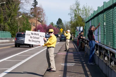 A determined activist in a bright yellow jacket and cap holds a handwritten protest sign reading "No Work No School No Shopping" while standing on North Skidmore Street's sidewalk above Interstate 5 in Portland. The late afternoon light casts long shadows across the concrete as fellow demonstrators gather along the green chain-link fence barrier, with spring foliage and residential buildings creating a suburban backdrop. Traffic flows steadily below as the Indivisible movement participants stage their May 1st General Strike awareness campaign.