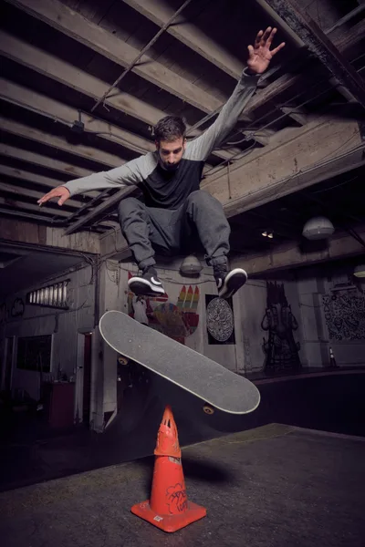 A bearded skateboarder launches into a gravity-defying ollie over a graffitied orange traffic cone atvCommonwealth Skateboarding in Portland's Buckman neighborhood. The moody lighting illuminates weathered wooden ceiling beams and concrete walls adorned with street art, while the athlete's arms spread wide in perfect aerial form. The underground venue's raw atmosphere creates a compelling contrast between urban grit and athletic grace.