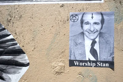 A provocative wheat paste poster adhered to a weathered concrete wall in Portland's Central Eastside features a black and white portrait of a smiling man in a suit and tie, adorned with occult symbols including a pentagram and inverted cross. The text "Worship Stan" appears beneath the portrait in bold white letters against a black banner. Adjacent torn paper fragments create textural contrast against the gritty urban surface, capturing the raw aesthetic of guerrilla street art in the Grand Avenue Historic District.