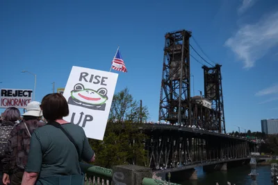 Demonstrators gather beneath Portland's iconic Steel Bridge during the March 2026 No Kings Protest, their signs and American flag silhouetted against a brilliant azure sky. A protester holds a distinctive 'RISE UP' placard featuring Pepe the Frog imagery, while the weathered steel lattework of the historic drawbridge looms overhead like an industrial cathedral. The scene captures a moment of civic engagement framed by the urban landscape of the Lloyd District, where political expression meets architectural heritage under the Pacific Northwest's crystalline light.