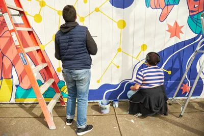 Two muralists collaborate on 'Let's Talk,' a vibrant community artwork at Open Signal in Portland's historic Eliot neighborhood. One artist, wearing a navy vest and jeans, observes the geometric yellow network pattern while their colleague in a blue striped shirt sits cross-legged on the concrete, headphones on, painting flowing blue and coral forms. An orange stepladder stands ready against the white brick wall, paint cans scattered nearby as the mural takes shape under natural daylight.