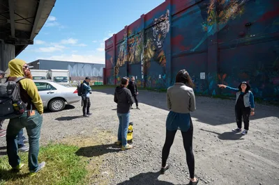 A diverse group of participants gathers on weathered asphalt beneath spring's clear blue sky, following tour guide Tiffany Conklin as she gestures toward vibrant murals adorning the industrial walls of Portland's Central Eastside. The scene captures the intersection of urban art and community engagement, where concrete infrastructure meets colorful creative expression. Shadows fall sharply across the gravel lot, while participants in casual attire crane their necks to absorb the cultural narratives painted across the warehouse facades.
