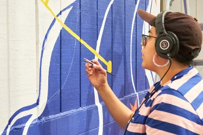 Maria Rodriguez, known as Sparkykneecap, works intently on a vibrant blue and yellow mural titled "Let's Talk" at Open Signal in Portland's Eliot neighborhood. Wearing headphones and a blue-striped shirt, the Mexican-American artist carefully applies paint to the wooden fence surface in bright afternoon light. The geometric composition captures the meditative focus of creation, with bold cerulean blues and sunny yellows forming abstract shapes that speak to themes of identity and cultural connection.