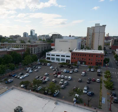 An elevated view captures the diverse architectural tapestry of Portland's Old Town district, where a surface parking lot serves as the immediate foreground beneath gathering afternoon clouds. The scene reveals the characteristic blend of historic red brick buildings and contemporary structures that define this Pacific Northwest neighborhood, with the Kalberer building prominently anchoring the middle ground. Soft natural light filters through a partly cloudy sky, casting gentle shadows across the urban landscape and creating depth between the foreground parking area and the distant high-rise towers.