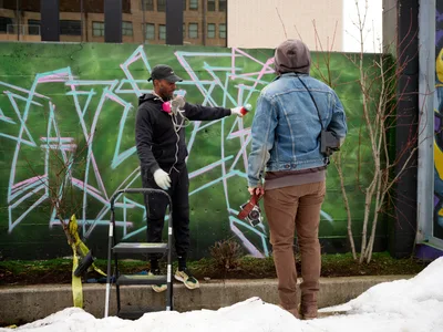 In the creative enclave of Southeast Portland's Hosford-Abernethy neighborhood, a street artist in black hoodie and cap applies spray paint to a vibrant geometric mural while engaging in conversation with a photographer in denim jacket and brown pants. The emerald green wall serves as canvas for angular white and pink lettering, with urban apartment buildings rising in the soft overcast light behind bare winter branches.