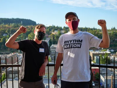 Michael Gaskill and Peter Marks raise their fists in solidarity against a sweeping vista of Oregon City's hillside neighborhoods, their masked faces reflecting the pandemic-era context of their climate activism. The afternoon light illuminates their determined expressions as they pose on the McLoughlin Promenade balcony, with the forested hills and residential sprawl of Clackamas County creating a layered backdrop. Gaskill wears a black shirt while Marks displays his commitment with a white "Rhythm Nation" tee, both men embodying the grassroots energy of environmental protest.
