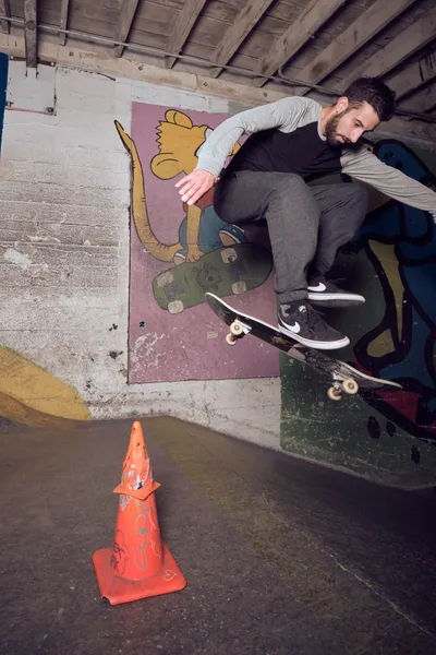 A bearded skateboarder in casual streetwear executes a wall ride against a vibrant mural in the raw concrete interior of Commonwealth Skateboarding in Portland's Buckman neighborhood. The industrial basement setting, marked by exposed wooden ceiling beams and weathered concrete walls, creates an authentic underground atmosphere. A spray-painted traffic cone sits prominently in the foreground, while colorful street art featuring a cartoon character adds urban energy to the gritty practice space.