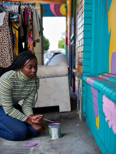 Naomi Likayi sits on the concrete sidewalk, carefully signing her vibrant turquoise and purple mural in Portland's Alberta Arts District. Wearing a striped green sweater, she holds a paintbrush over an open paint can, adding her signature to the colorful geometric artwork that adorns the building's exterior wall. The scene captures an intimate moment of artistic completion against the backdrop of Northeast Portland's creative corridor, with clothing displays and urban street life visible through the shop's interior.