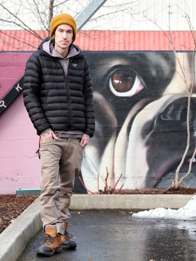 Muralist Wetiko stands confidently before his completed street art piece at the Clay Creative Building in Southeast Portland's Hosford-Abernethy neighborhood. Dressed in winter layers with a distinctive mustard beanie and black down jacket, the artist poses against his striking mural featuring an oversized eye rendered in dramatic blacks and earth tones. The late winter setting captures bare branches overhead and patches of melting snow, while the industrial creative space provides an authentic backdrop for this portrait of contemporary urban artistry.