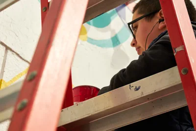 Artist Anke Gladnick works intently on their mural commission "Let's Talk" within the creative spaces of Open Signal in Portland's Eliot neighborhood. Framed by coral-pink scaffolding and surrounded by paint supplies, the illustrator embodies focused artistic dedication as natural light illuminates their workspace. The scene captures the intersection of public art creation and community cultural programming in Northeast Portland's vibrant arts district.