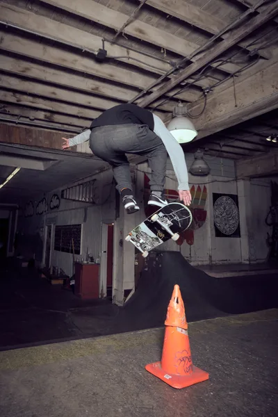 A skateboarder executes an airborne trick over a weathered orange traffic cone in the industrial interior of Commonwealth Skateboarding in Portland's Buckman neighborhood. The raw concrete floors and exposed ceiling beams create a gritty urban atmosphere, while graffiti and street art adorn the walls beneath harsh industrial lighting. The skater, captured mid-flight in dark clothing, demonstrates technical skill against the backdrop of this authentic underground skate venue.