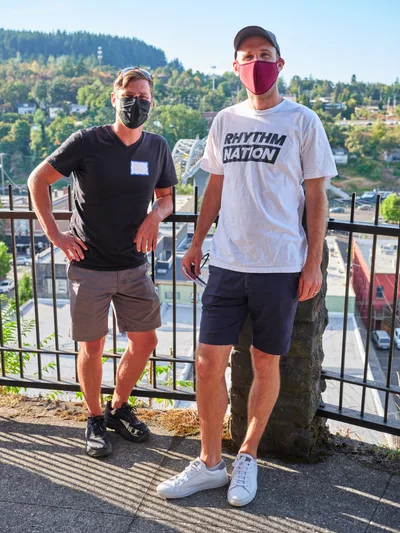 Michael Gaskill and Peter Marks stand side by side on McLoughlin Promenade overlooking Oregon City, both wearing protective masks during a climate change protest at Representative Kurt Schrader's office. The golden hour light casts dramatic shadows through metal railings as the forested hills and residential neighborhoods stretch into the distance. Gaskill wears a black v-neck with khaki shorts while Marks sports a white "Rhythm Nation" t-shirt and navy shorts, their relaxed poses contrasting with the serious political nature of their gathering.