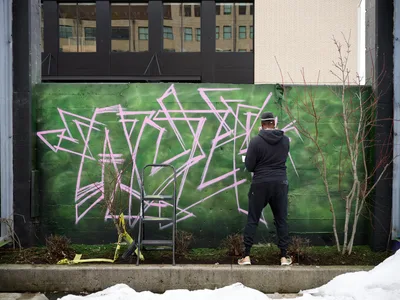 A hooded artist works methodically on an elaborate pink and white graffiti piece against a vibrant green wall in Southeast Portland's Hosford-Abernethy neighborhood. The scene unfolds in Clay Creative's parking lot, where winter-bare saplings frame the emerging artwork while urban architecture provides a backdrop of glass and brick. Yellow caution tape and construction materials hint at the ongoing transformation of this creative space.
