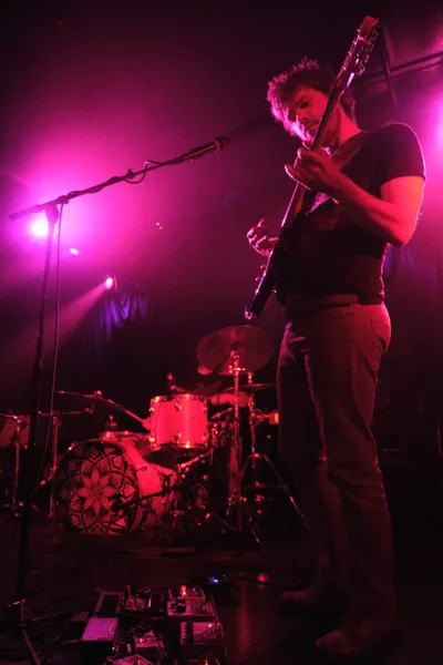 A guitarist from Sun Blood Stories commands the stage at Dante's in downtown Portland, bathed in deep magenta and red stage lighting that creates an otherworldly atmosphere. The musician, dressed in casual dark clothing, grips an electric guitar while positioned near the microphone stand, with a drum kit visible in the atmospheric haze behind him. The intimate venue setting captures the raw energy of live music performance, where dramatic lighting transforms the mundane into the cinematic.