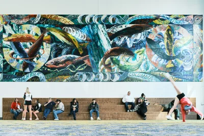 Visitors seek momentary refuge on stone benches beneath an explosive panorama of maritime-themed murals at Portland's Oregon Convention Center. The vibrant wall art, dominated by swirling blues and oceanic motifs interwoven with text fragments, creates a dramatic backdrop for the diverse group of attendees—from a breakdancer frozen mid-move to couples and individuals in casual repose. The juxtaposition of kinetic street art energy above and human stillness below captures the convention center's role as both cultural crossroads and temporary sanctuary.