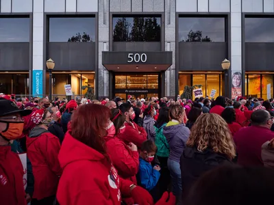 A dense crowd of healthcare workers, predominantly wearing red Kaiser Permanente scrubs and winter jackets, gathers in solidarity outside the Kaiser Permanente Tower at 500 Northeast Multnomah Street in Portland's Lloyd District. The sea of crimson creates a powerful visual statement against the modern glass and steel facade of the medical facility, while protest signs punctuate the crowd and evening light casts a determined atmosphere over the assembled nurses. Face masks worn by many participants underscore the contemporary healthcare context of their labor action.