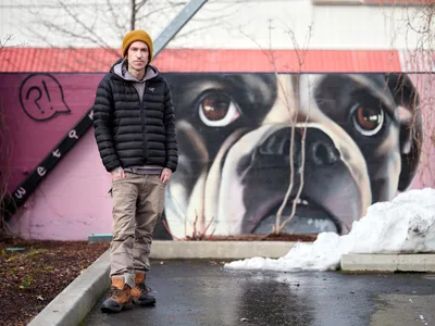 Painter and muralist Wetiko stands confidently before his striking dog portrait mural at Clay Creative Building in Southeast Portland's Hosford-Abernethy neighborhood. The artist wears a golden mustard beanie and black puffer jacket against the late winter atmosphere, with patches of snow visible on the concrete. Behind him, his hyperrealistic mural depicts a close-up of a dog's face with expressive brown eyes and detailed fur work, painted in warm tones against the building's coral-pink exterior wall.