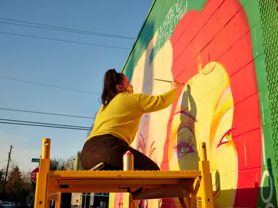 Multidisciplinary artist Villatose works from a wooden scaffolding platform, adding delicate brushstrokes to her large-scale mural 'Jardín Cubana' depicting three female figures on Northeast Alberta Street in Portland's celebrated arts district. Golden hour light bathes the scene, illuminating her mustard yellow sweater against the mural's bold palette of coral, emerald, and sunshine yellow. Power lines stretch across the azure sky above, while spray paint cans rest on the platform beside her as she brings life to the colorful portrait emerging on the building's facade.