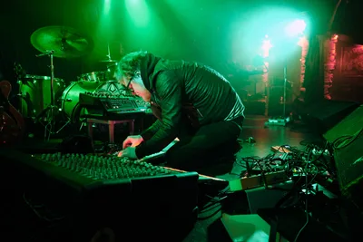 Electronic musician Eli Savage, performing as ellipsism, hunches intensely over a mixing console amid a sea of synthesizers and equipment at Portland's legendary Dante's venue. Ethereal green stage lighting bathes the intimate performance space, creating an otherworldly atmosphere as the artist manipulates sound through an array of electronic instruments. Behind him, a drum kit and additional gear fade into the moody shadows, while tangled cables snake across the stage floor, evidence of the complex sonic architecture being constructed in real time.