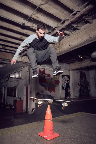 A bearded skateboarder in grey sweats and black tee defies gravity in the raw concrete underbelly of Commonwealth Skateboarding in Portland's Buckman neighborhood. The industrial space's exposed wooden ceiling beams frame this moment of athletic suspension as he soars above a weathered orange traffic cone, his skateboard floating beneath him. Graffiti-tagged walls and shadowy recesses create an authentic urban backdrop for this editorial skateboarding portrait.