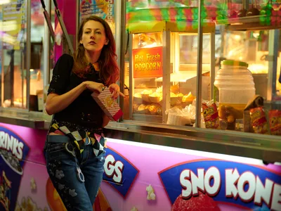 A young woman in distressed denim and a sequined black top leans against a vibrant concession stand at the Clark County Fair in Vancouver, Washington. The golden glow from the popcorn machine illuminates her contemplative expression as neon carnival lights create a dreamy bokeh in the background. The scene captures the nostalgic magic of fairground evenings, where the aroma of kettle corn mingles with the electric energy of carnival attractions.