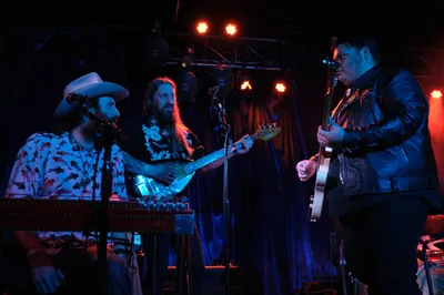 Barry Walker in a cowboy hat and floral shirt commands the keyboards while Tobias Berblinger and Victor Franco flank him with guitar and bass in the atmospheric lighting of Dante's club. Crimson stage lights pierce through the intimate venue's darkness, casting the Portland trio in warm amber and electric blue hues as they deliver their performance during RX Fest 2019. The cramped stage setup and close quarters create an electric intimacy between the musicians and their audience.