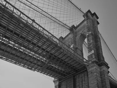 A dramatic worm's-eye view captures the Gothic Revival stonework of the Brooklyn Bridge's tower through the intricate steel lattice of the bridge deck structure above. Shot in monochrome from Emily Warren Roebling Plaza in DUMBO, the composition emphasizes the engineering marvel's dual nature - the romantic masonry arches juxtaposed against the industrial precision of steel cables and support beams. The low angle transforms the familiar landmark into an abstract study of intersecting lines and geometric patterns, with the tower's crenellated crown piercing the overcast Brooklyn sky.
