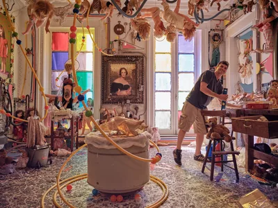 Artist Mikel Glass navigates through his fantastical installation space at The Cell Theatre on West 23rd Street, surrounded by an explosion of suspended dolls, colorful geometric window panels, and eclectic found objects. The scene pulses with creative energy as Glass moves through this immersive wonderland of theatrical props and artistic debris. Natural light filters through the rainbow-hued window panes, casting jewel-toned shadows across the cluttered floor where vintage portraits and whimsical toys create a dreamlike tableau. The atmosphere captures the raw, unfiltered moment before an artistic transformation unfolds.