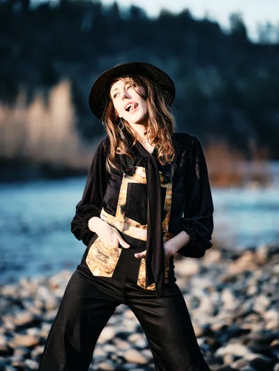 A young woman radiates pure joy as golden hour light illuminates her face along the rocky riverbed of Oregon's Floodplain Trail in Multnomah County. She wears a black wide-brimmed hat and layered earth-toned clothing, her mouth open in mid-laugh as she poses playfully against the backdrop of the Columbia River and distant forested hills. The warm, directional sunlight creates dramatic contrast between her sunlit figure and the soft blue tones of the water and sky behind her.