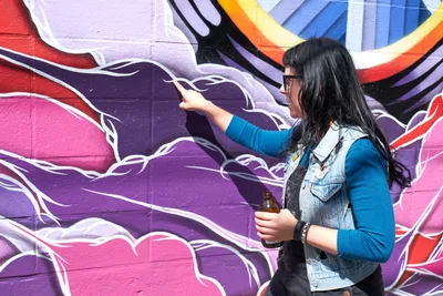 Tiffany Conklin of the Portland Street Art Alliance gestures toward a vibrant purple and pink mural while leading an interpretive tour in Portland's Central Eastside neighborhood. Dressed in a blue sweater and denim vest, she holds a bottle while explaining the artwork's significance against the backdrop of flowing, organic forms painted in deep purples, magentas, and warm orange accents. The scene captures the intimate, educational nature of community-led cultural tours.
