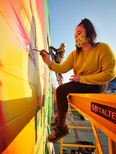 Multidisciplinary artist Vilatose applies vibrant brushstrokes to 'Jardín Cubana,' a mural depicting three female figures, while perched on an orange Metaltech scissor lift against the warm golden hour sky. The scene captures creative collaboration as another artist works in the background on the concrete wall. Wearing a patterned face mask and mustard yellow sweater, the artist embodies the resilient spirit of Portland's Alberta Arts District, where public art transforms urban landscapes into canvases of community expression.