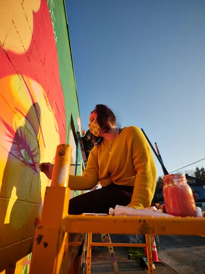 Multidisciplinary artist Villatose works from an orange scissor lift against a cloudless blue sky, painting the bold mural 'Jardín Cubana' featuring three female figures in tropical coral and yellow hues on Northeast Alberta Street. The masked artist, wearing a golden yellow knit sweater, concentrates on their work while surrounded by paintbrushes and materials in the heart of Portland's Alberta Arts District. The golden hour light bathes the scene in warm tones, highlighting the contrast between the vivid wall art and the serene evening atmosphere.