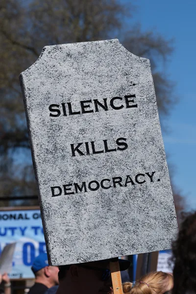 A weathered granite-textured protest sign shaped like a tombstone bears the stark message "SILENCE KILLS DEMOCRACY" in bold black lettering against Portland's azure winter sky. The foam board memorial, held aloft during the March 2026 No Kings demonstration in Old Town's historic Skidmore district, looms above glimpses of other protesters and campaign signage below. Bare tree branches frame the scene, creating a somber backdrop that amplifies the sign's grave warning about civic participation.