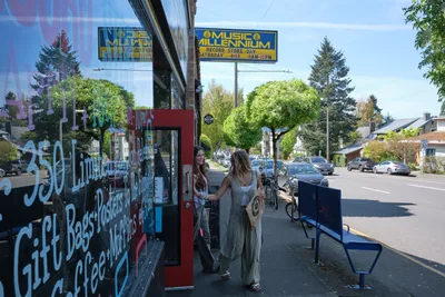 Two young women browse vintage records outside Music Millennium, Portland's iconic record store on East Burnside Street. The afternoon light casts gentle shadows across the sidewalk as one woman in a flowing dress examines vinyl while her companion watches from the doorway. The scene captures the enduring appeal of physical music media against the backdrop of Portland's eclectic Kerns neighborhood, where colorful storefronts and tree-lined streets create an authentic urban village atmosphere.