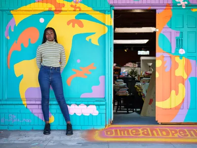 Naomi Likayi stands confidently beside her vibrant abstract mural on Northeast 15th Avenue in Portland's Alberta district. The young Black woman, dressed in a striped turtleneck and dark jeans, poses against the explosion of turquoise, yellow, coral, and lavender forms that transform the building's facade. The mural's organic shapes and playful palette create a striking contrast with the urban streetscape, while the open doorway reveals the interior of what appears to be a community space or shop.