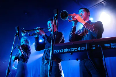 Two trumpet players from the band Roselit Bone perform in dramatic blue stage lighting at Dante's in Portland, Oregon during RX Fest 2019. The musicians, dressed in dark clothing, stand behind a Nord Electro 4 HP keyboard, their brass instruments gleaming under the moody concert illumination. Microphones capture their performance while intense backlighting creates an atmospheric haze, emphasizing the intimate venue setting of this underground music festival.