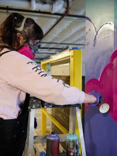 A focused artist in protective mask and gloves spray-paints vibrant purple and magenta forms onto a wall within the industrial confines of Clay Creative's parking garage in Southeast Portland. The scene captures the intersection of urban art and creative workspace, with aluminum ladders and spray cans arranged around the emerging mural. Warm artificial lighting illuminates the work in progress, creating an intimate documentation of street art creation in Portland's Hosford-Abernethy neighborhood.