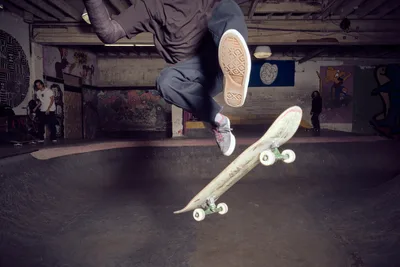 A skateboarder's legs and board are captured mid-air above the curved concrete of Commonwealth Skateboarding's bowl in Portland's Buckman neighborhood. The atmospheric lighting illuminates weathered sneaker soles and the deck's underside while graffiti-covered walls and fellow skaters create an authentic underground scene. The low-angle perspective emphasizes the height and technical precision of the aerial maneuver.