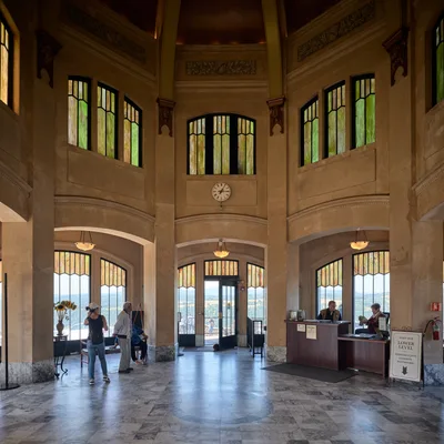 The soaring interior of Vista House reveals its 1918 architectural grandeur, with honey-toned stone walls rising to an octagonal crown adorned with stained glass windows. Visitors move quietly through the formal space, their footsteps echoing across polished marble floors as natural light filters through amber and green glass panels. An information desk anchors the ceremonial hall, while the central clock marks time beneath ornate carved details that speak to the structure's role as both scenic overlook and architectural monument.