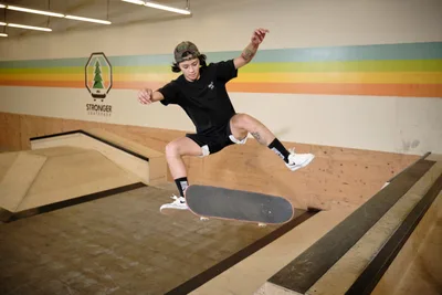 Professional skateboarder Christiana Means executes a dynamic aerial maneuver above the wooden bowl at Stronger Skatepark in Milwaukie, Oregon. Suspended in mid-flight with arms outstretched for balance, she demonstrates precise board control against the venue's distinctive rainbow-striped wall mural. The warm fluorescent lighting illuminates the polished plywood surfaces and creates an intimate atmosphere within this Pacific Northwest skateboarding sanctuary.